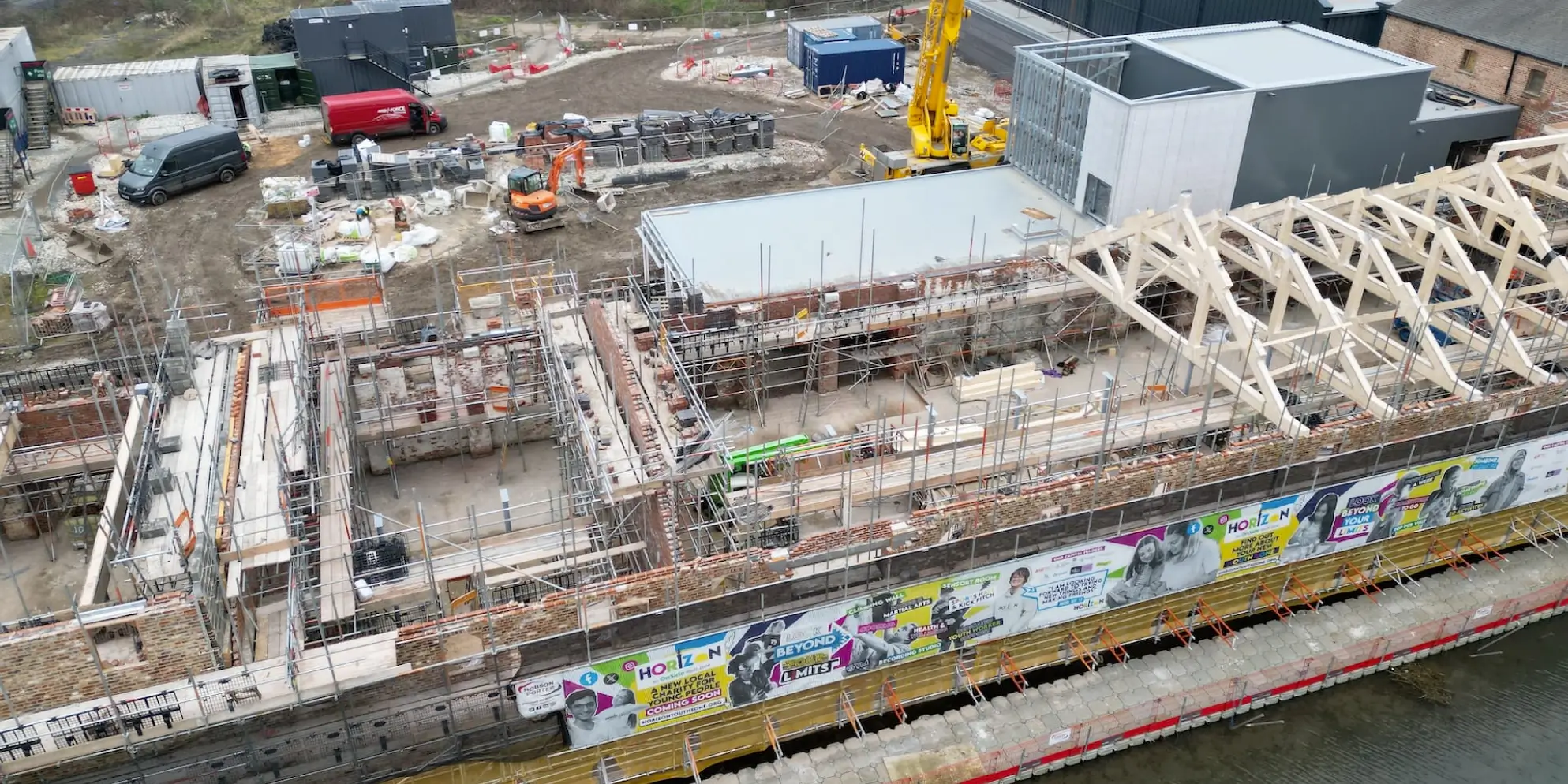 At the construction site, partially built structures rise alongside a large yellow crane, signaling contributions to Grimsby's Future. Nearby, assorted building materials and machinery surround a red vehicle and orange equipment, setting the foundation for the Horizon Youth Zone.